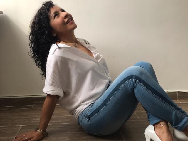 Etoile, con cabello negro rizado, camisa blanca y jeans azules, sentada en un piso de madera, sonriendo, reclinada hacia atrás con las manos en el suelo, usando zapatos planos blancos y una pulsera dorada..