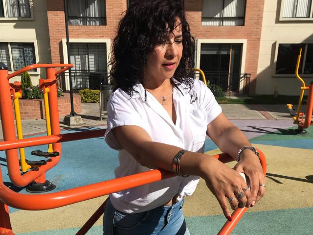 EtoileLatin with curly black hair, white shirt, blue jeans, leaning on orange playground bars, wearing bracelets and rings, sunlight on her face, smiling slightly, outside apartment building.