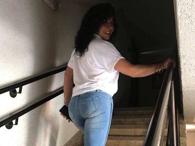 EtoileLatin with curly black hair, white shirt, and tight blue jeans, smiling while holding a black phone, standing on stairs, showing her round ass.