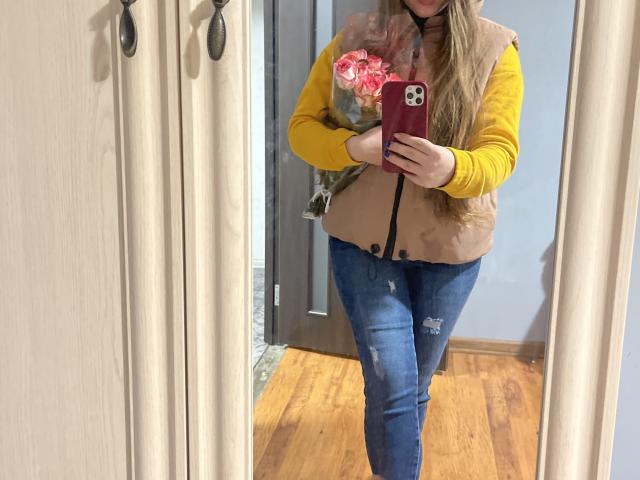 Dooris with long blonde hair, yellow shirt, brown vest, blue jeans, holding pink flowers, taking a mirror selfie with a red phone, wooden floor, beige curtains, and a door.