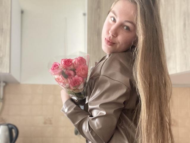 Dooris with long, straight brown hair, holding pink roses, wearing a brown leather jacket, looking over her shoulder with a subtle smile, in a kitchen.