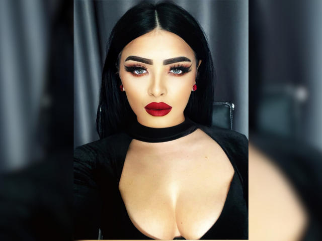Devasha, com cabelo preto, batom vermelho e maquiagem pesada nos olhos, vestindo um colar preto e uma blusa preta com decote profundo que mostra seus seios. Seus brincos vermelhos combinam com seu batom..