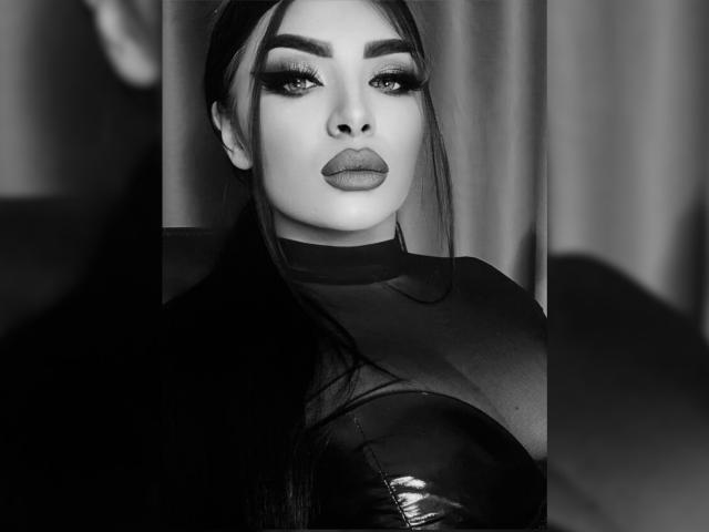 Black and white photo of Devasha with full eyebrows, dark lipstick, and heavy eyeliner. She has long dark hair, a sheer top, and a shiny black top.