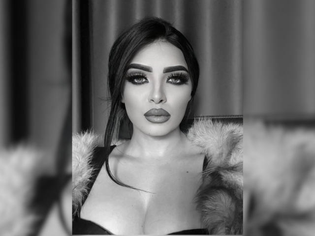 Black-and-white photo of Devasha with dark, slicked-back hair, heavy eyeliner, and full lips. She's wearing a furry coat, showing off her large, round tits.