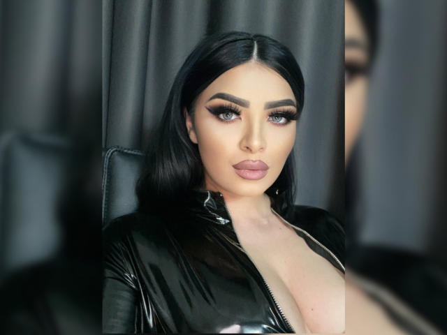 Devasha with long black hair, heavy eyeliner, and pink lipstick, wearing a shiny black zip-up top, exposing her large tits. She's sitting against a dark background.