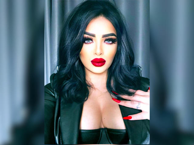 Devasha with black hair, red lipstick, and dramatic eye makeup, wearing a black leather top, showing large tits, and red nails, standing in front of gray curtains.