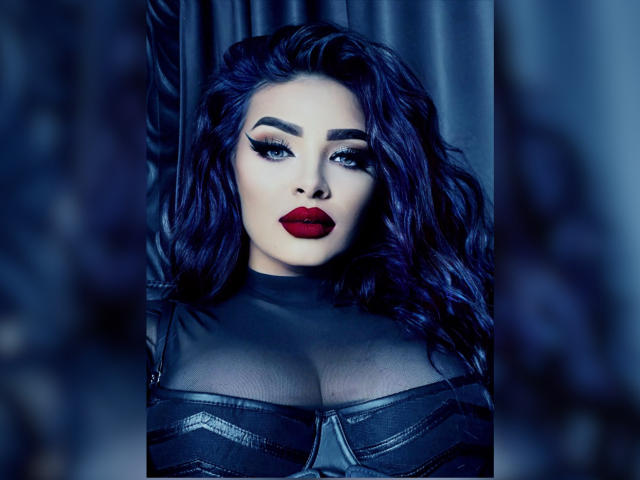 Devasha with dark blue wavy hair, red lipstick, and black leather outfit, showing large boobs, looking seductively at the camera in a moody, blue-lit photograph.