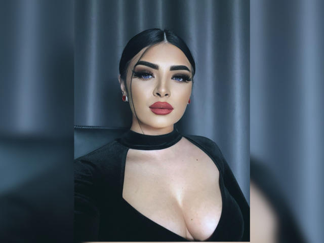 Devasha with black hair, heavy eyeliner, and red lipstick, wearing a black dress with a deep cleavage, showing her large tits, sitting against a grey curtain.