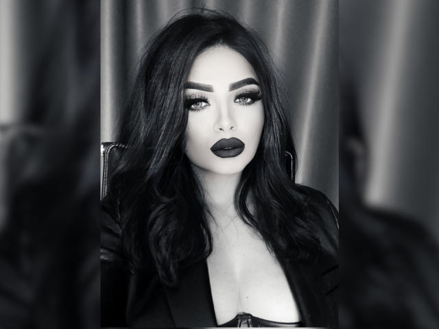 Black-and-white photo of Devasha with dark, wavy hair, heavy makeup, and bold lipstick, sitting, showing cleavage in a low-cut top, with a serious, seductive expression.