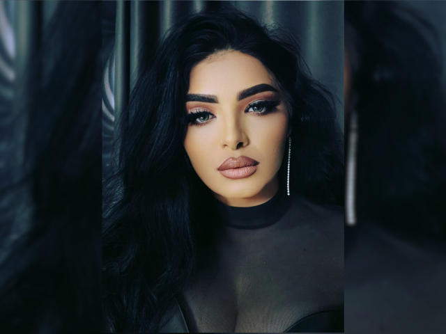 Devasha with long black hair, full lips, and dramatic eye makeup. She's wearing a sheer black top, showing a little cleavage, with a long silver earring.