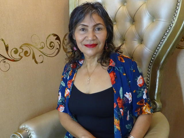 CelesteBakers has curly brown hair, red lipstick, and wears a blue floral shirt over a black top, sitting on a fancy, silver-tufted chair, smiling. Her tits are small.