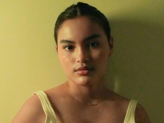 MiiaPalmer has shiny, slightly sweaty skin, brown eyes, and dark hair pulled back. She wears a white tank top with thin straps and a delicate necklace.