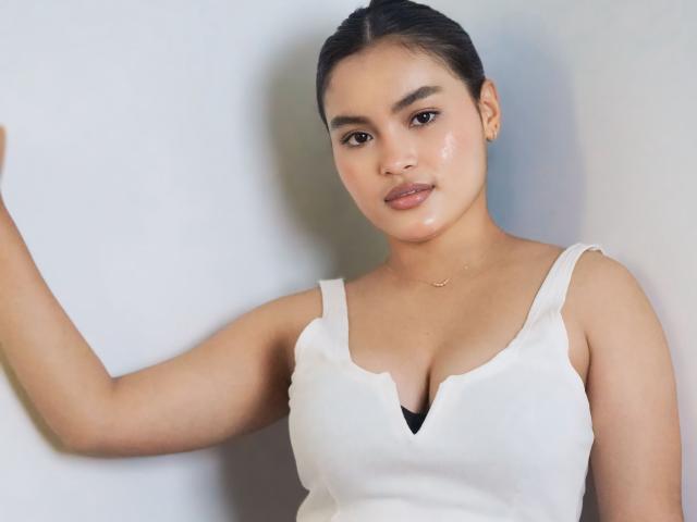 MiiaPalmer with medium tits, wearing a white tank top, black bra peeking, short dark hair, light skin, subtle makeup, gold necklace, standing against a plain wall, looking at camera.
