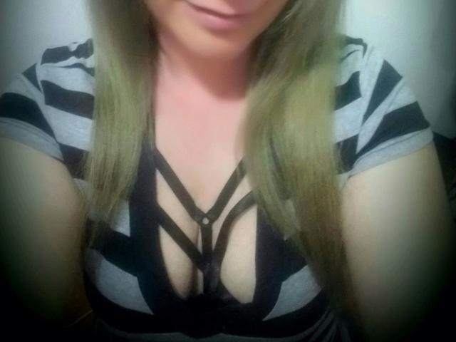 SecretTemptationX with blonde hair, striped grey and black shirt, and black strappy bra, showing her big tits and cleavage. Soft, slightly blurry selfie.
