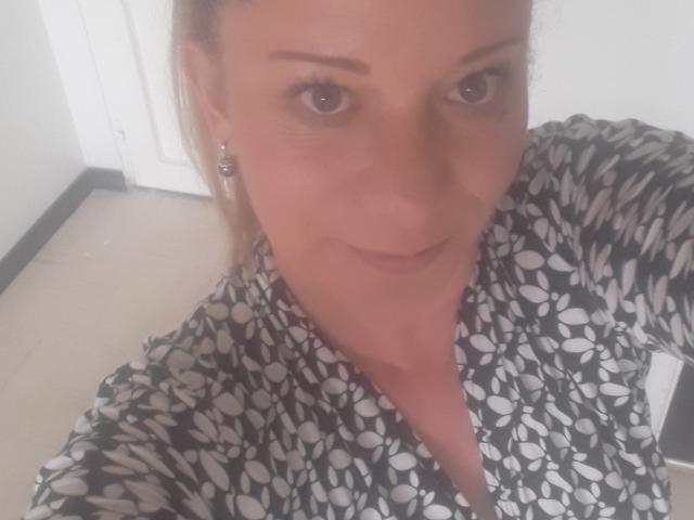 SecretTemptationX with light brown hair, wearing a black and white floral shirt, has subtle makeup, small earrings, and a slight smile, looking into the camera.