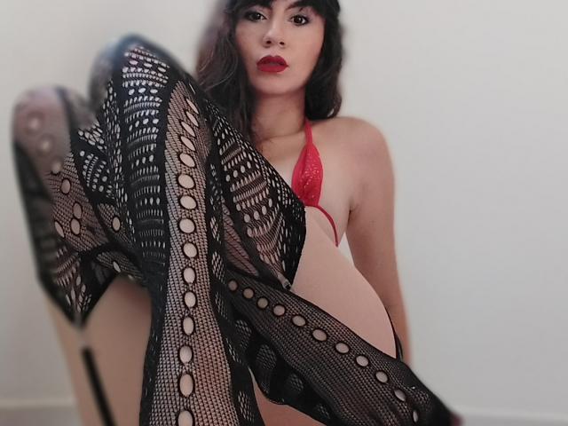 Kari Carter indossa un top da bikini rosso, calze a rete nere e rossetto rosso, mostrando i piedi in primo piano, seduta con le gambe sollevate. Ha i capelli scuri e un aspetto seducente..