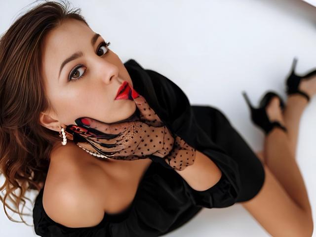 SweetBB69 with red lipstick, black polka-dot gloves, and black dress, lying on white surface, wearing black heels, hand on cheek, looking up, brown wavy hair, red nails.
