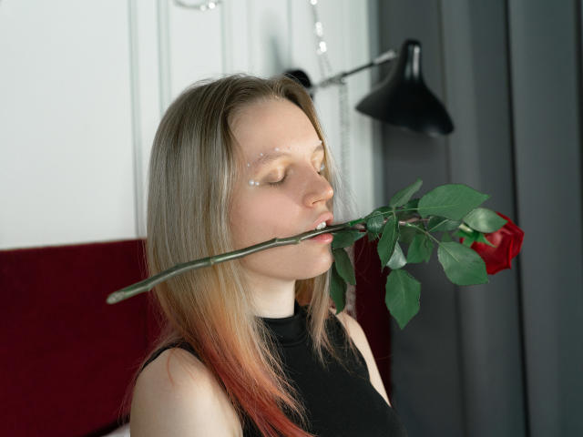 Beatrica Klein, con i capelli biondi, che tiene una rosa rossa in bocca, indossa una canotta nera senza maniche, occhi chiusi, trucco scintillante e leggero, una lampada nera sullo sfondo, luce naturale soffusa..