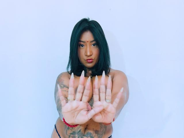 Nahirr with black hair, tattoos, and long nails, hands out, showing off her tits, wearing a red bracelet, looking at the camera with pink lipstick.
