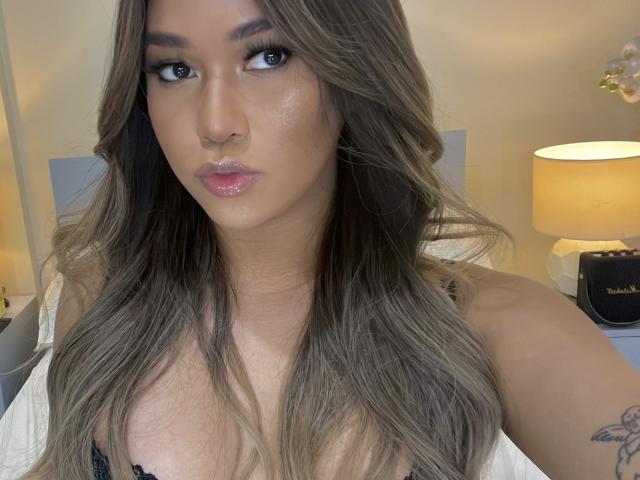 MissTati has long, wavy brown hair, shiny pink lips, a nose ring, and a tattoo on her right arm. She's wearing a black top, showing her tits.