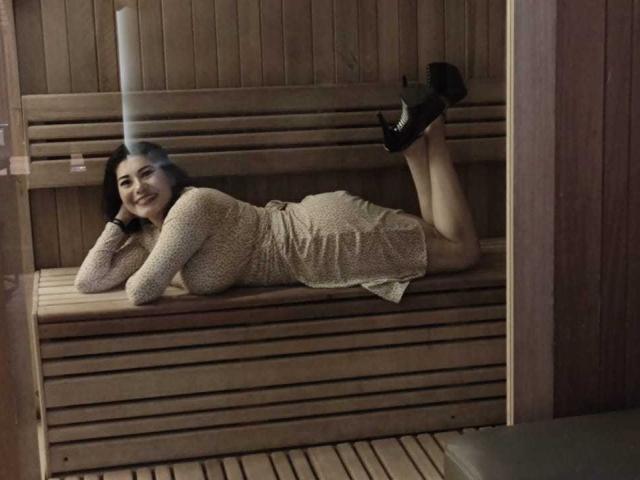 InsaciableIntensa, lying on wooden bench, wearing beige knit dress, black high heels, smiling, arms under chin, legs bent at knees, wooden walls, relaxed, cute, attractive.