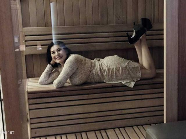 InsaciableIntensa, lying on wooden bench, wearing a beige, long-sleeve, textured dress, black high heels, smiling, hands under chin, legs bent, ass slightly raised, wooden walls.
