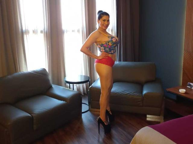 InsaciableIntensa stands in a room, wearing a floral top, red lace panties, and black high heels, with hands on hips, showing off her tits and ass.