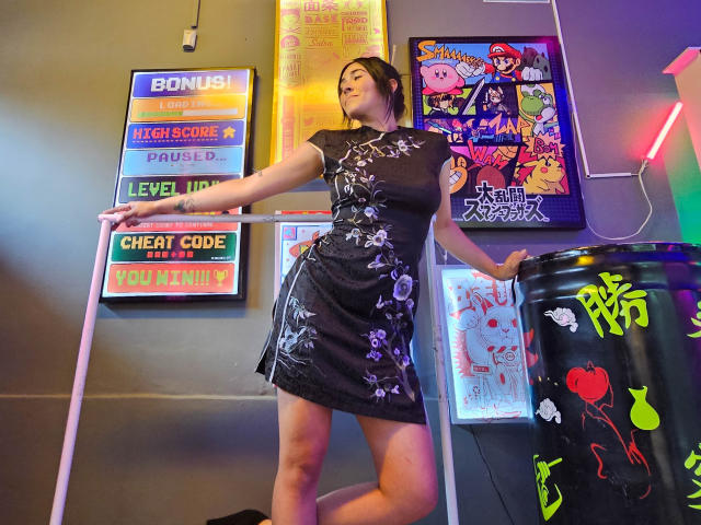 NicoColeman in a black floral dress, standing with one hand on a railing, her ass slightly raised, surrounded by colorful posters, neon lights, and a black trash can.