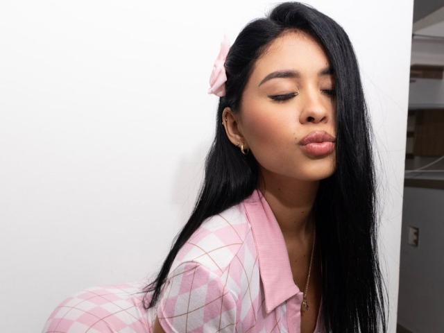 AddaBella, con i suoi lunghi capelli neri, l'outfit a quadretti rosa e il fiocco rosa, con le labbra rosa leggermente schiacciate, indossa orecchini dorati, con gli occhi chiusi, davanti a un muro bianco..