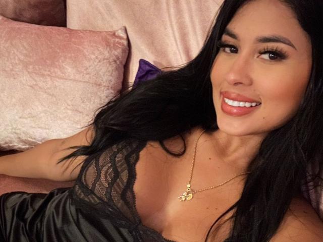 AddaBella, con i capelli scuri, sorride, indossa una maglia di pizzo nero che mostra il seno, una collana d'oro, è sdraiata su cuscini di velluto rosa, ha la pelle abbronzata, labbra piene ed è rivolta verso la fotocamera..