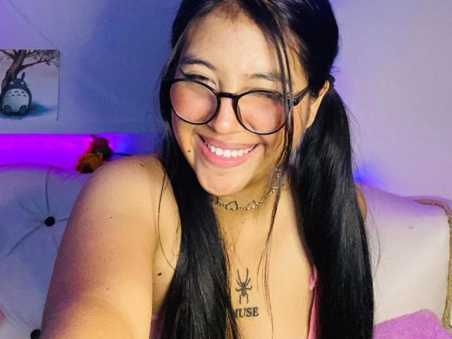 Elizaa mit schwarzen Zöpfen, lächelnd mit geschlossenen Augen, trägt eine Brille, eine Halskette und ein Spinnentattoo unter ihrer Brust. Ihr pinker BH ist sichtbar..