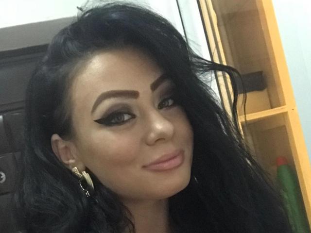 MaysAT has dark, wavy hair, bold eyebrows, and thick black eyeliner. She's wearing gold hoop earrings and has a slight, confident smile with pink lipstick.