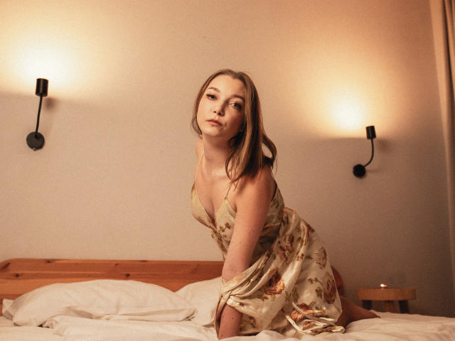 LovelyxEmilly on a bed, wearing a floral silk dress, leaning forward with her tits slightly visible, blonde hair, wall sconces, wooden headboard, white pillows, and a wooden nightstand.