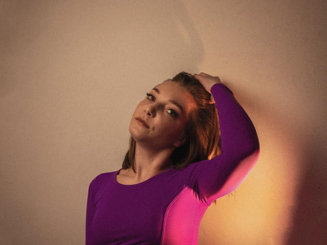 LovelyxEmilly, une main dans ses cheveux bruns, portant un haut à manches longues violet et rose, regardant vers le haut avec un léger sourire, une ombre douce sur son visage..
