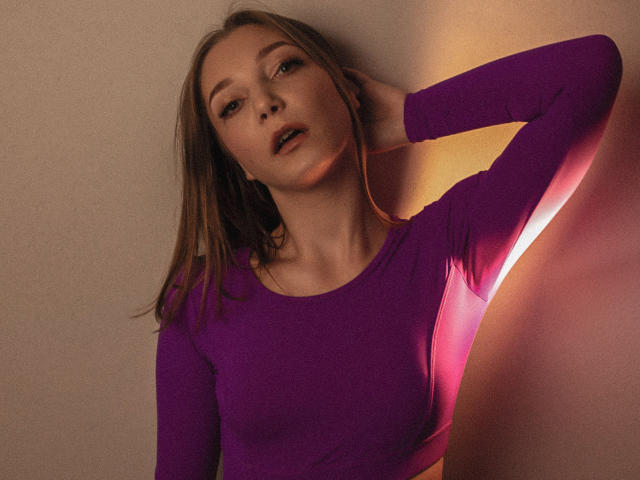 LovelyxEmilly, avec ses longs cheveux bruns, portant un haut violet moulant, a une main dans ses cheveux, et regarde légèrement vers le haut avec une expression douce, sous un éclairage chaud et rosé..