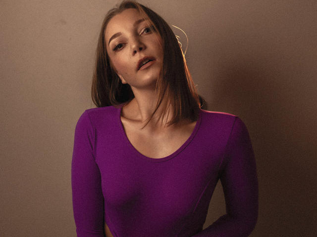 LovelyxEmilly, avec une poitrine de taille moyenne, portant un haut violet moulant à manches longues, des cheveux bruns, un léger maquillage sur le visage, regardant en dehors de la caméra avec une expression détendue. Éclairage doux et chaleureux..