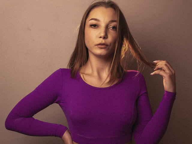 LovelyxEmilly with brown hair, purple long-sleeve shirt, holding hair with right hand, serious expression, light skin, small tits, hand on hip, neutral brown background, soft lighting.
