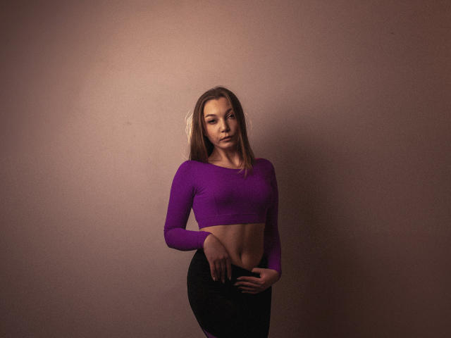 LovelyxEmilly, con cabello largo y castaño, vestida con un top corto morado y pantalones negros, de pie frente a una pared beige, mostrando un abdomen delgado y plano, y una pose segura y sensual..