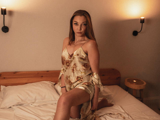 LovelyxEmilly met lang bruin haar, gekleed in een bloemenprint zijden jurk, zittend op een bed met witte kussens, waarbij haar achterwerk gedeeltelijk zichtbaar is, kijkend naar de camera met een serieuze uitdrukking..
