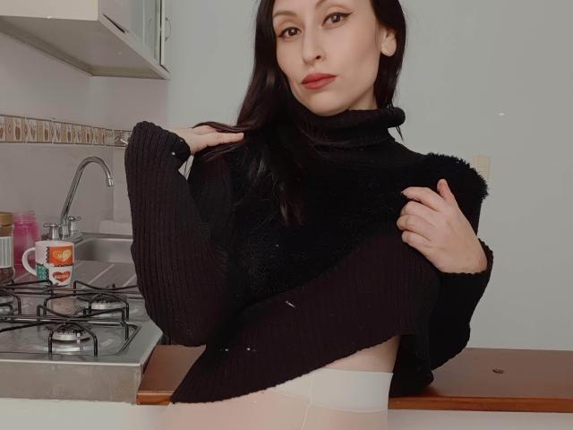 AliceRossi with long dark hair, wearing a black turtleneck, lifting her sweater to reveal white panties, standing in a kitchen, raising one leg, showing her smooth, bare ass.