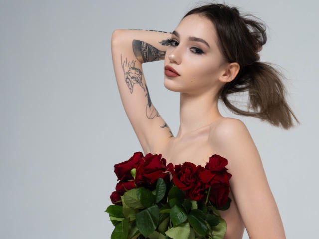 SophieKitty with red roses covering her tits, arm raised, showing tattoos, brown poneytail, light skin, subtle makeup, sexy, confident, gray background.