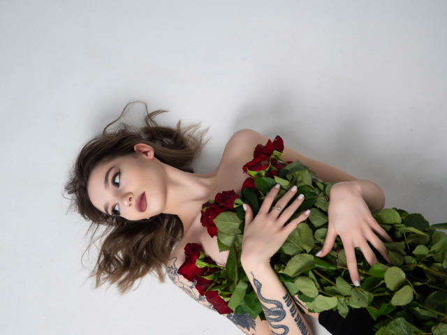 SophieKitty with red roses, holding them against her tits, looking up with a pensive expression, tattoos on her arm, lying on a white surface, brown wavy hair.