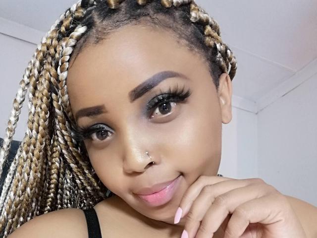 TangyYummyAss has light brown skin, braided blonde and black hair, black eyeliner, long eyelashes, a nose piercing, and a cute smile with pink lips, resting her chin on her hand.
