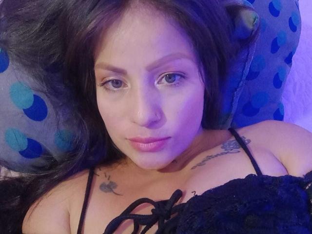Angela, acostada, con cabello negro, un piercing en la nariz y tatuajes en el pecho, vestida con una blusa negra de encaje, mostrando sus pequeños senos, y mirando a la cámara con una luz púrpura..