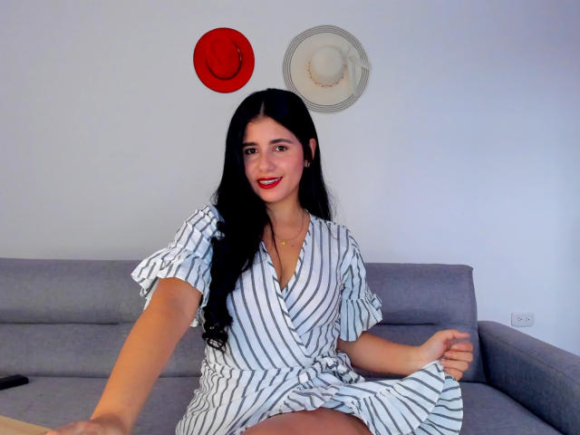 NatashaSwtst has long black hair, red lipstick, and is wearing a striped dress, sitting on a gray couch, with red and white hats on the wall behind her.