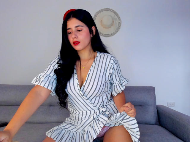 NatashaSwtst with long black hair, red lipstick, and striped dress, sitting on gray sofa, pulling up dress to reveal pink panties, small tits, cute, sexy, white wall in background.