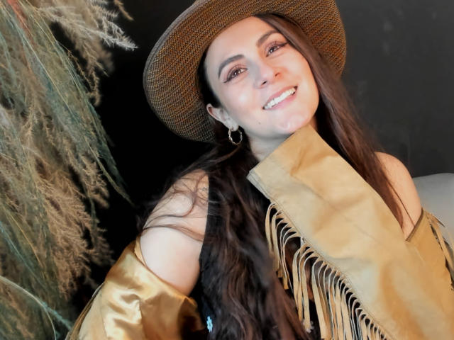 StephanieMarrien with long brown hair, smiling, wearing a tan hat, gold earrings, and a fringed tan shirt. Her tits are partially covered by the shirt. Her ass is not visible.