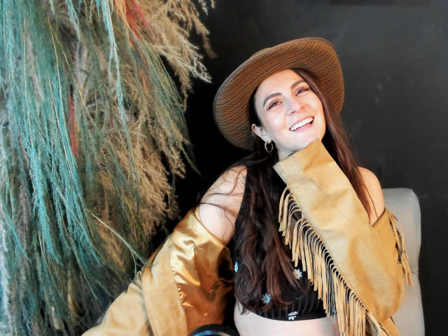 StephanieMarrien with long brown hair, smiling, wearing a brown cowboy hat, tan fringed jacket, and hoop earrings, has tits partially covered by the jacket, standing next to greenery.