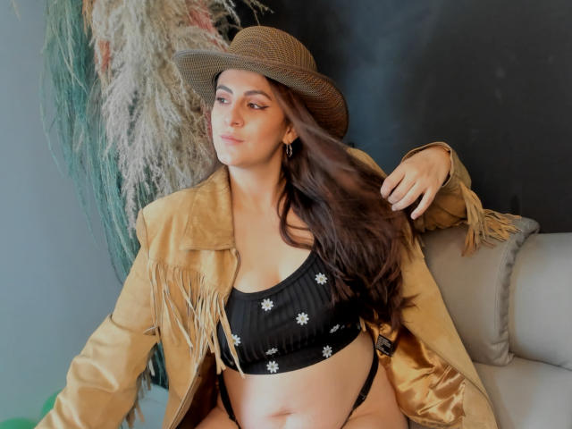 StephanieMarrien with brown hat, tan fringe jacket, black flower bra, and black thong, showing her tits and pussy, sitting on a couch, long brown hair, earrings.