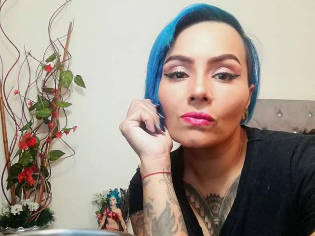 AbbyCrown mit blauen Haaren, Tätowierungen, schwarzem T-Shirt, pinkem Lippenstift, Eyeliner im "Winged"-Stil, Hand am Kinn, Blick in die Kamera, rotes Armband, rote Blumen im Hintergrund, leicht schmollender Gesichtsausdruck..